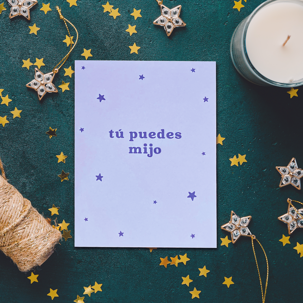 "Tú puedes mijo" purple greeting card on an emerald green background surrounded by gold star confetti, a white candle, some star ornaments, and yarn. 