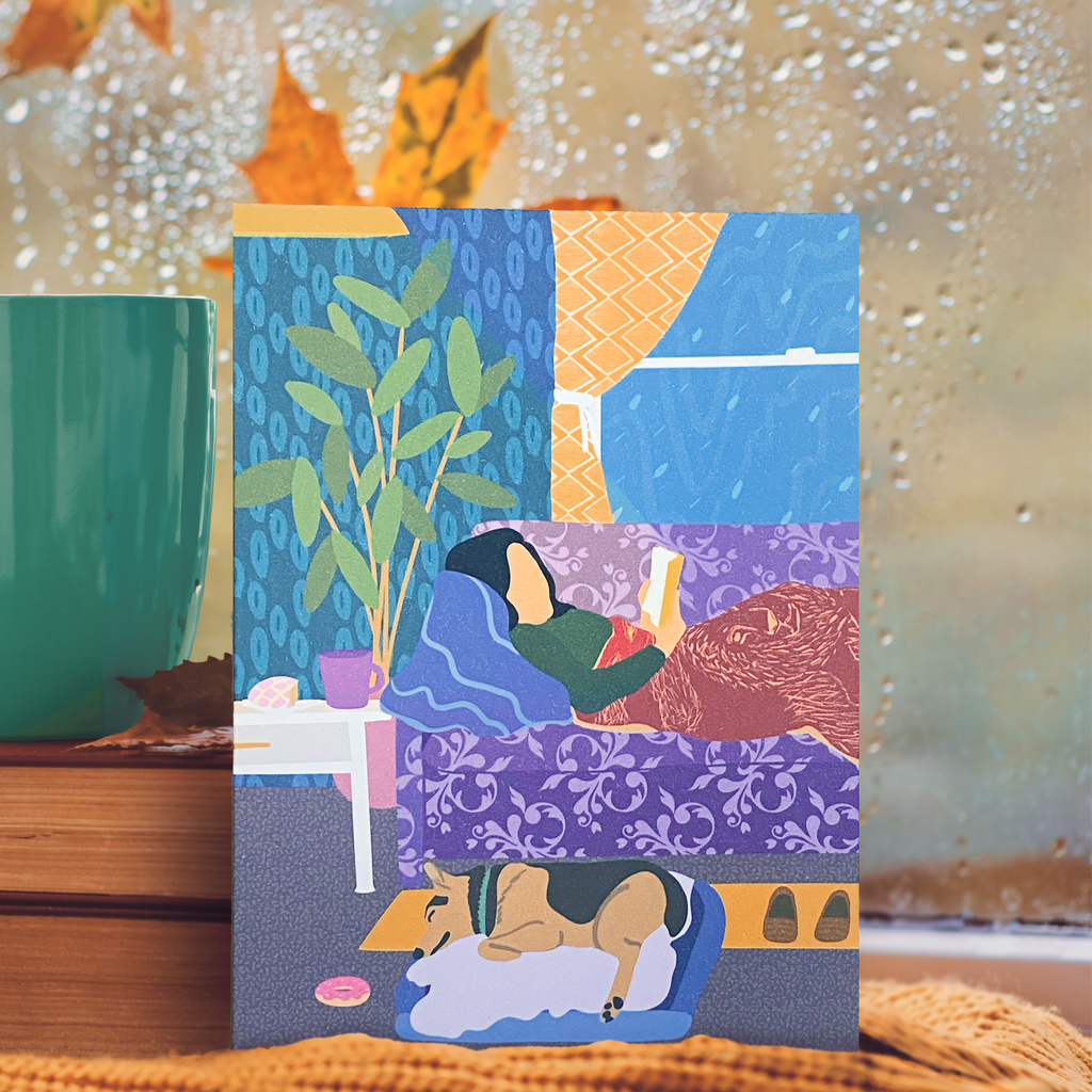 "Cobija Weather" greeting card featuring a woman lying down on a couch reading a book while covered with a red lion design blanket.  Her dog is sleeping in its bed next to her on a rainy day. 