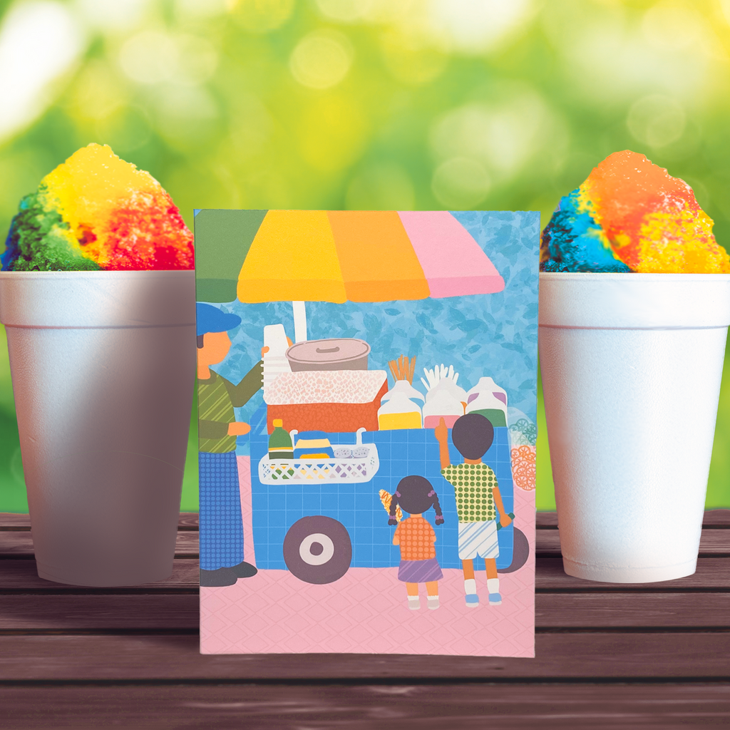 Raspados greeting card product photo.  Greeting card features two children in front of a cart where a person is selling elotes, raspados, and chicharrones.  The card is displayed on a table with a multi-colored raspado on each side.