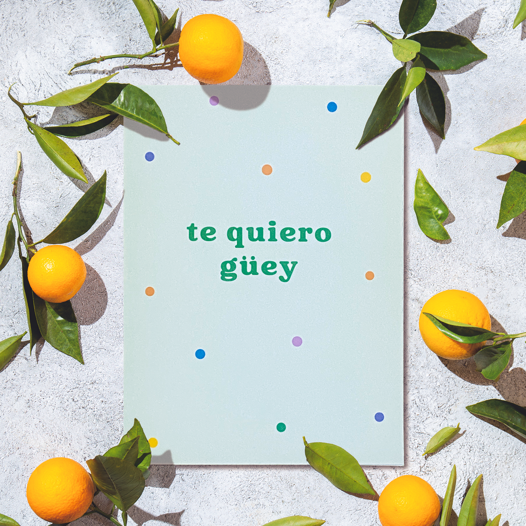 Portrait style greeting card with the Spanish phrase "te quiero güey" in dark green lowercase letters on a light green background and multi-colored dots around it. Card is on a background of cement with small oranges decorating the area around the card.