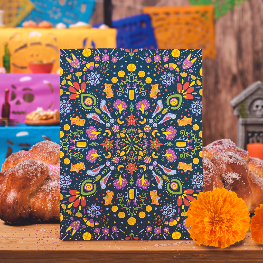 Dia de los muertos themed portrait style greeting card with a pattern that repeats radially on a deep blue background. The design features bright orange pan de muerto and puerquitos, cempasuchil flowers, and even a purple xoloitzcuintli dog among other graphics. The card is displayed upright on a table with pan de muerto behind it and a marigold in front of it.